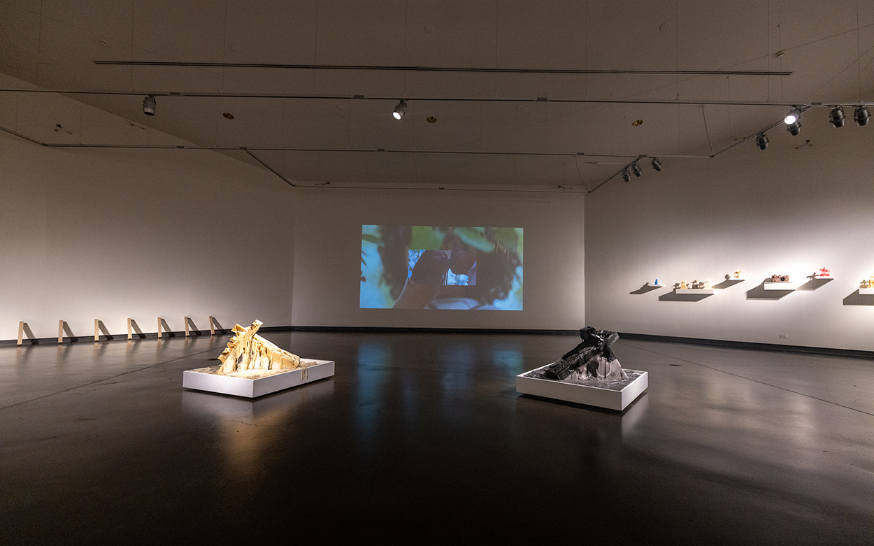 A sparsely appointed gallery floor displays two sculptures while images from a multimedia piece are projected on a wall in the background