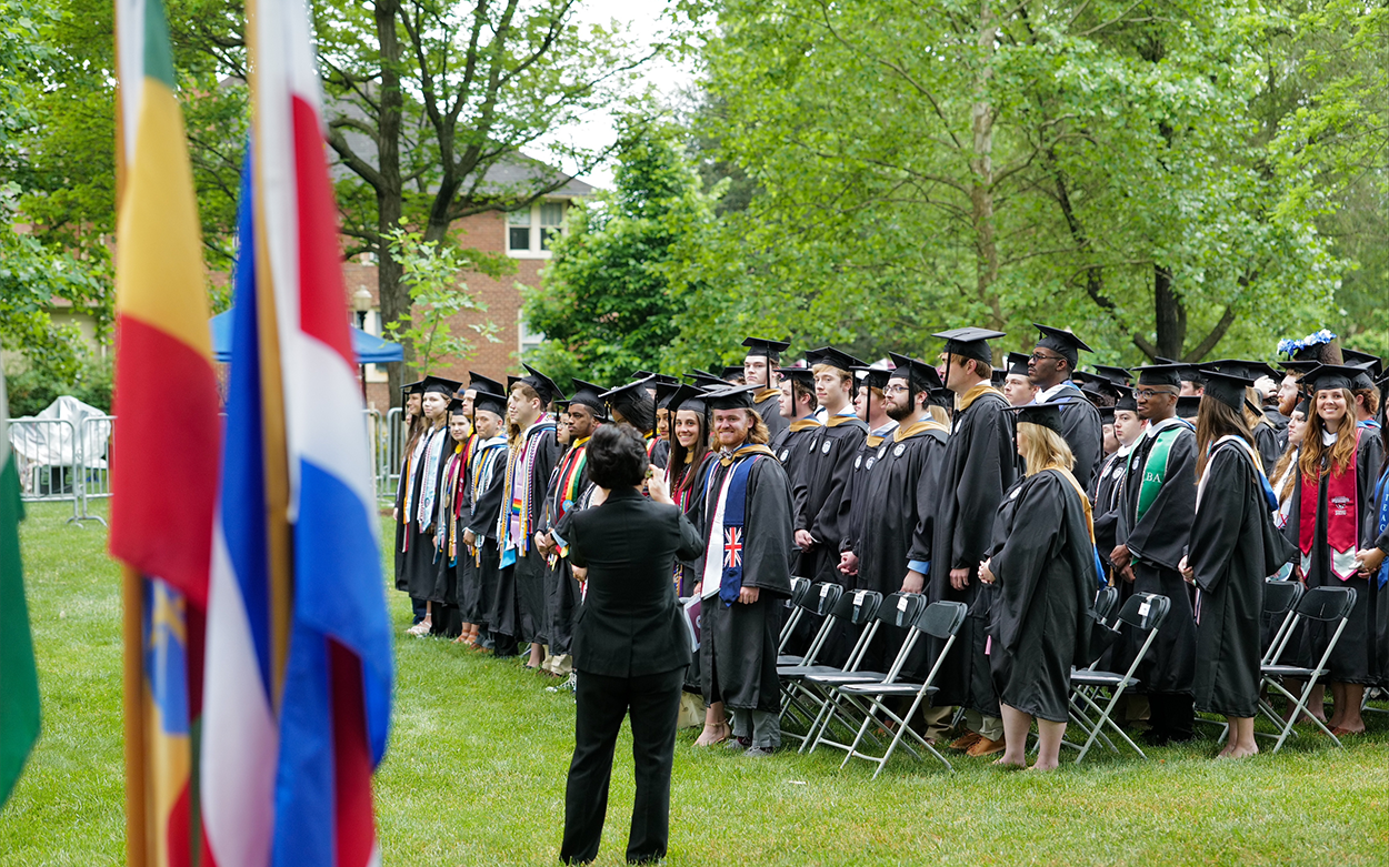 colorful flags and rows of students in graduation gowns with one person taking a photo