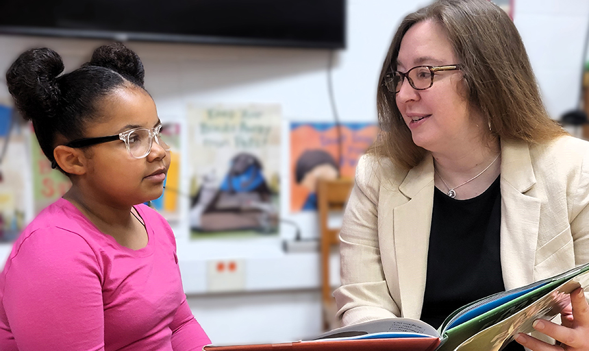 Woman in white blazer reads to a little girl with pigtails and a pink shirt.