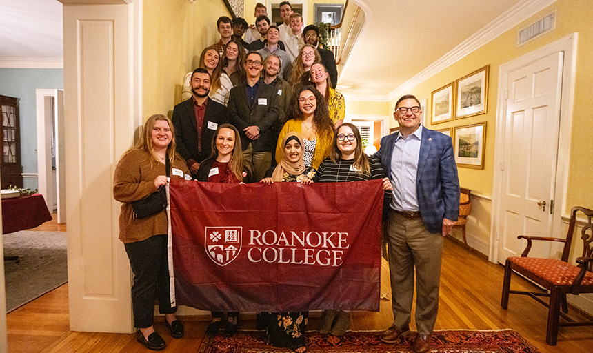 A group of young men and women stand on a staircase inside a large home. They are holding a big maroon flag that says Roanoke College.