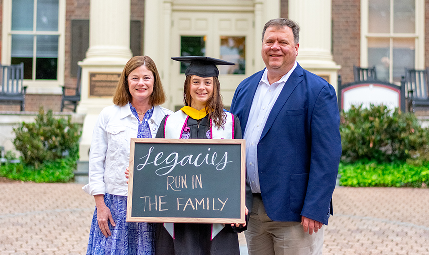 Mom and dad stand with their daughter between them, and daughter is wearing graduation attire and holding a chalkboard sign that says "Legacies run in the family."