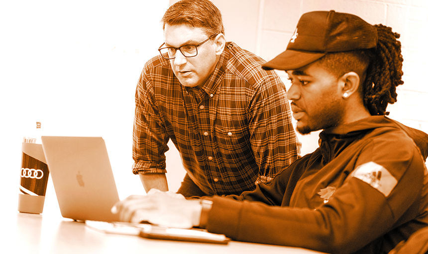 Orange-tinted photo of a student in a ball cap looking at a laptop while a professor looks over his shoulder.