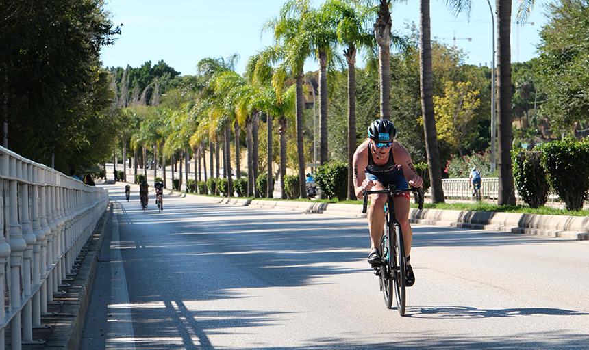 A woman cycles down a four-lane highway lined with palm trees. In the distance behind her are other cyclists.