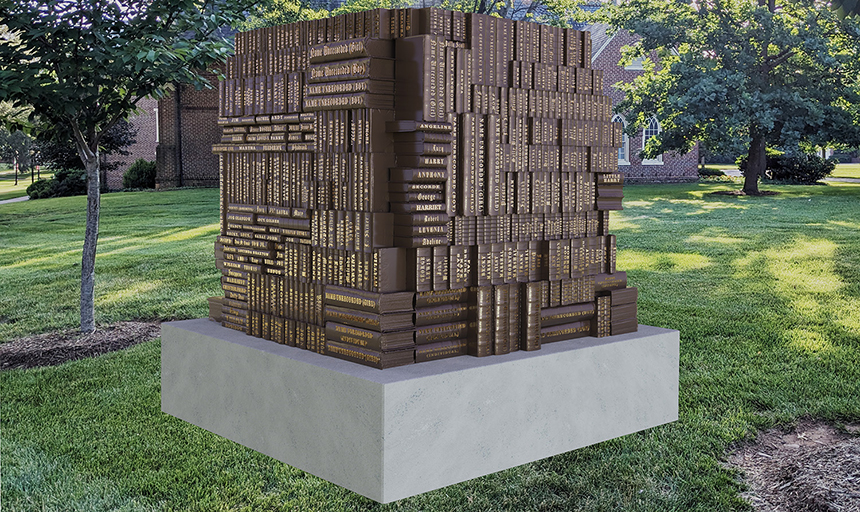 Square-shaped culpture consisting of stacks of bronze books, each of which has a name in gold lettering on the spine. It sits on a concrete or marble pedestal in a grassy landscape with a brick building behind it.