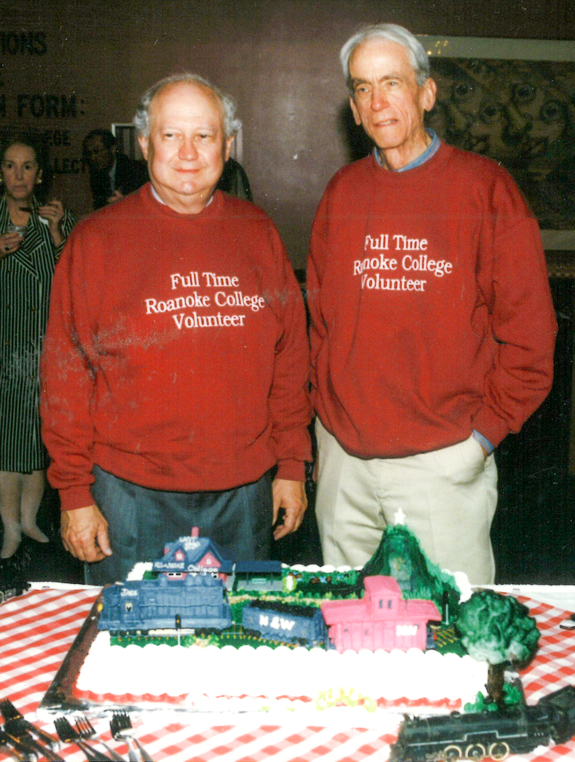 Two men stand matching red sweatshirts that say "Full Time Roanoke College Volunteer." They are standing in front of a cake with a train on it. 