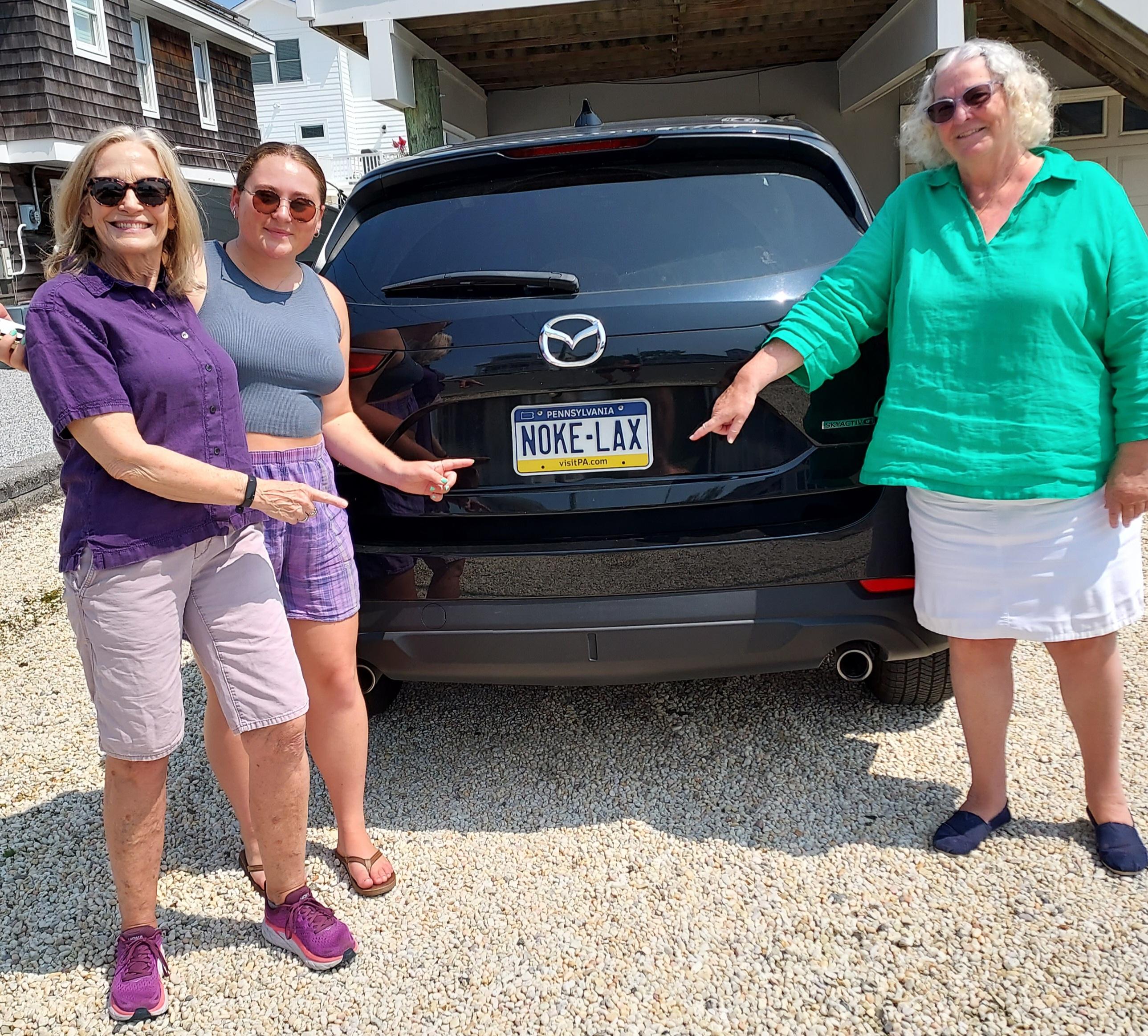 Three women stand at the rear of a sport utility vehicle and point at the license plate, which reads NOKE-LAX