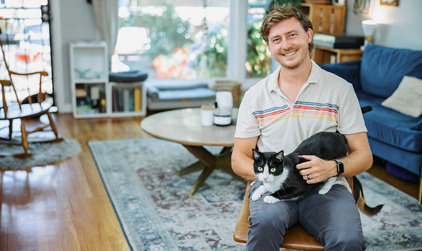 Man in short-sleeved white polo sits in a chair in a well-lighted room with a black and white cat on his lap.