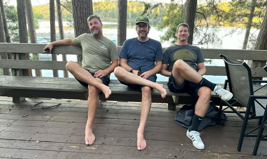 Three men sit on a bench with a lake in the background. They are all wearing shorts and t-shirts, and two are barefoot.