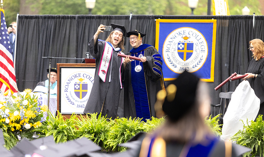 Young man in black robe pauses for a selfie with the college president as he crosses the graduation stage.