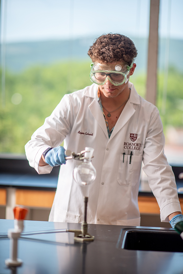 Student in a white lab coat with curly hair holds a beaker over a heat source