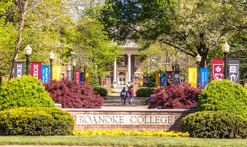 Campus view with green trees, Roanoke College flags in yellow, blue, black and red, and a wall that says "Roanoke College" across it.