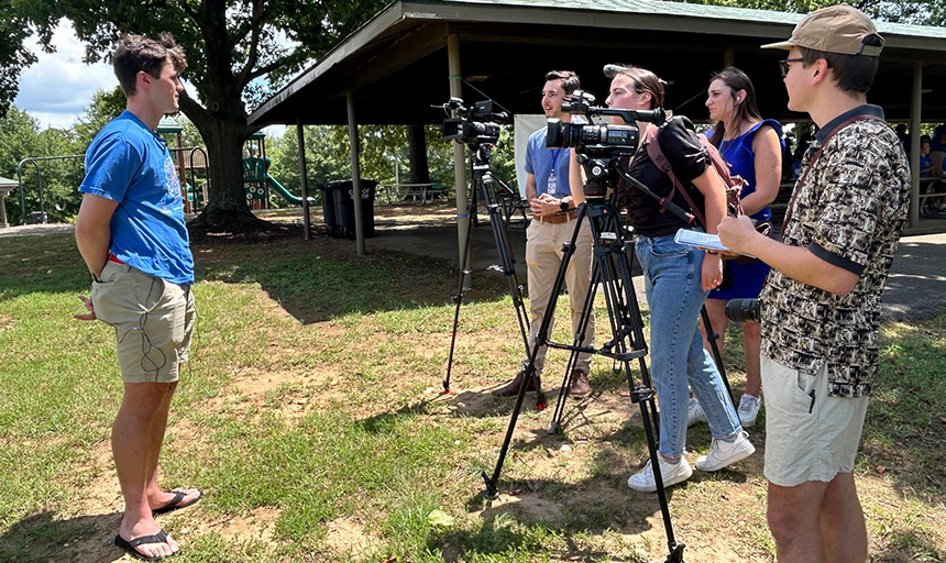 Young man is interviewed by three people with news cameras and one man holding a notebook.