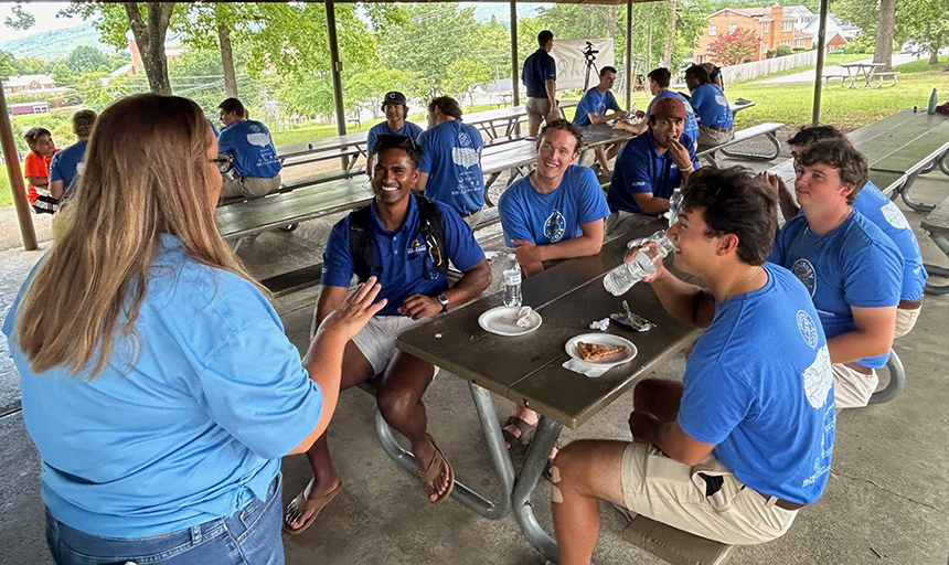 Woman stands at head of picnic table chatting with young men, who are sitting at the table eating pizza.