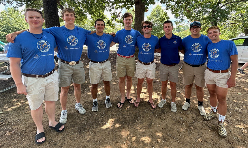 Eight young men in blue t-shirts and tan shorts stand in a row with their arms around each other smiling, with trees in the background