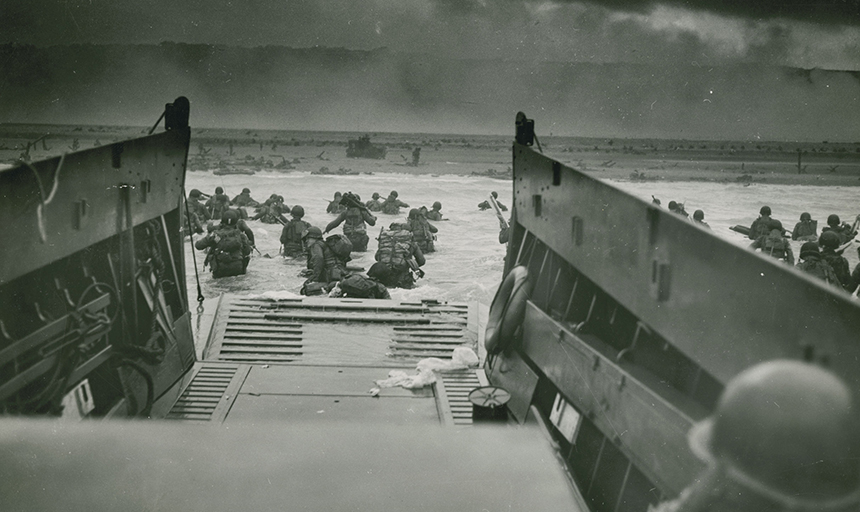 Servicemen pour out of a landing craft vehicle and wade through the ocean toward the French coastline. Mines, other watercraft and men can be seen in the distance.
