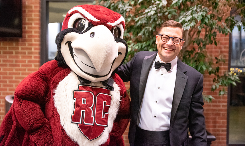 Man in a black tuxedo with his arm around a person in a mascot costume. Mascot is a maroon bird with RC on its chest.