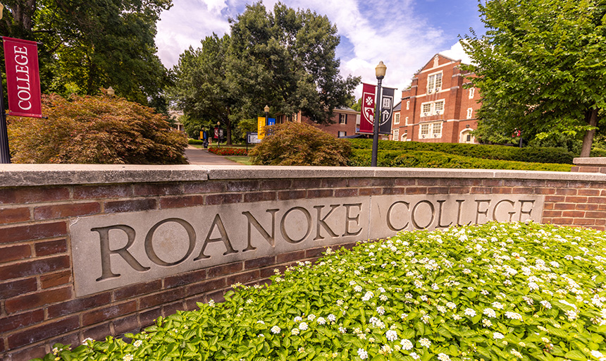 Brick wall with "Roanoke College" set in concrete and white flowers in foreground.