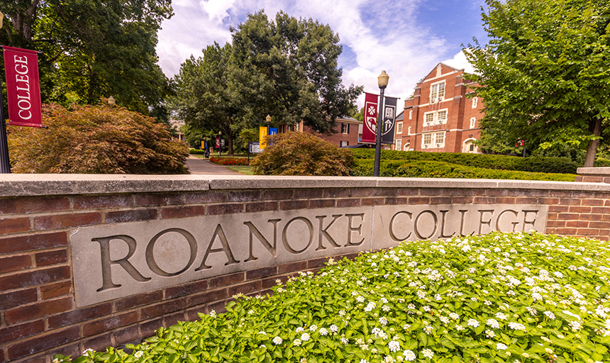 Brick and concrete Roanoke College sign with white flowers blooming in the foreground and campus buildings and trees in the background.