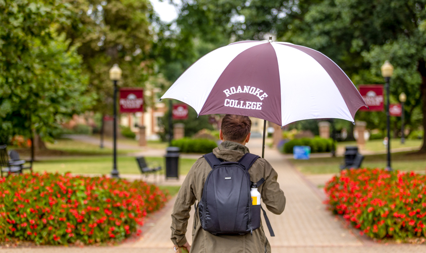 Man walking down a brick path holding a large white and maroon Roanoke College umbrella