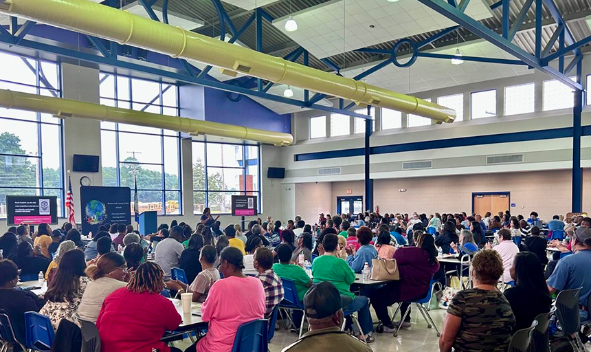 People gathered around tables in a school cafeteria listen to a training presentation