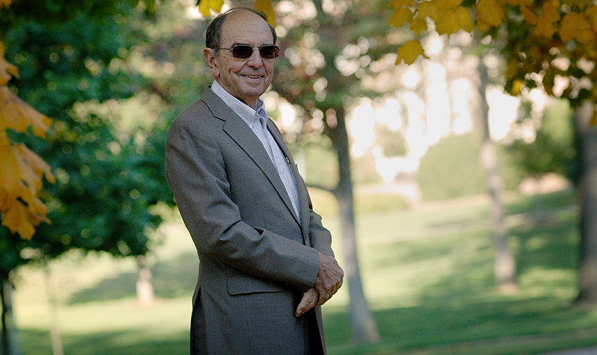 Portrait of an older man in a gray suit standing in a grassy quad with trees in the background