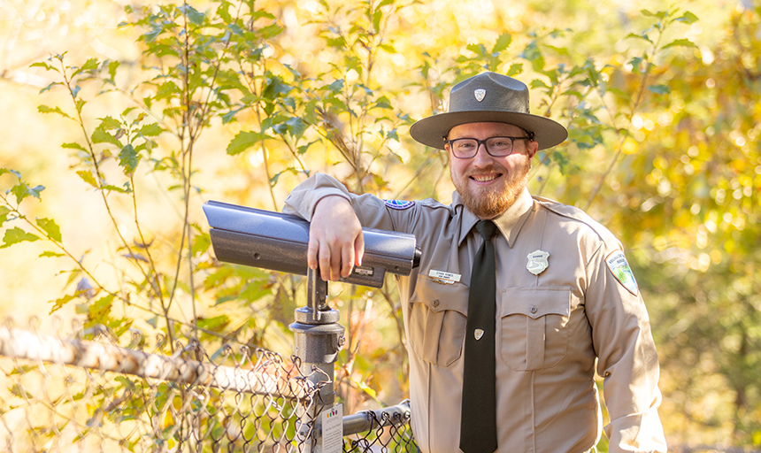 Man in a park ranger uniform leans against a viewfinder that allows colorblind park visitors to see in full color.