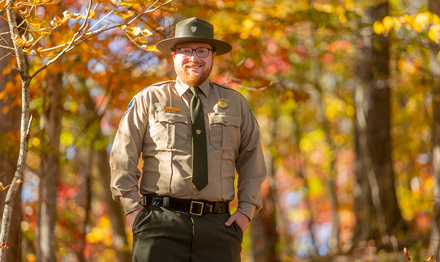 Man in a park ranger uniform and hat standing in front of a forest backdrop in autumn colors of orange and yellow.