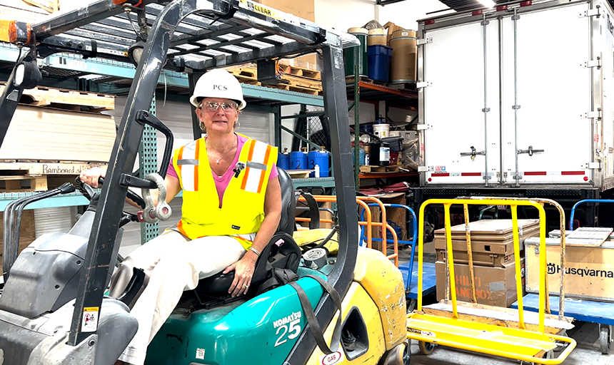 Woman in a bright yellow vest and white hard hat sits behind the wheel of a piece of heavy equipment.