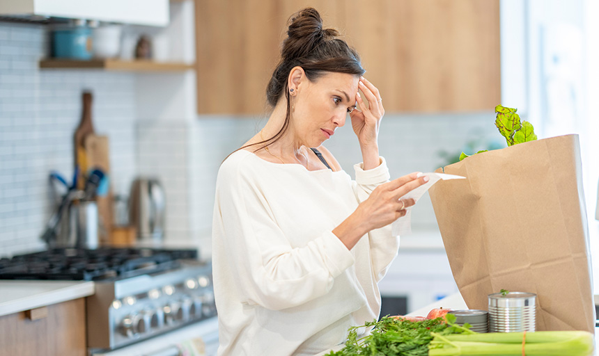 Woman in a kitchen examines a grocery receipt with a worried expression