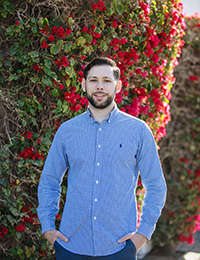 Young man in blue button-down shirt standing in front of a bush loaded with red flowers.