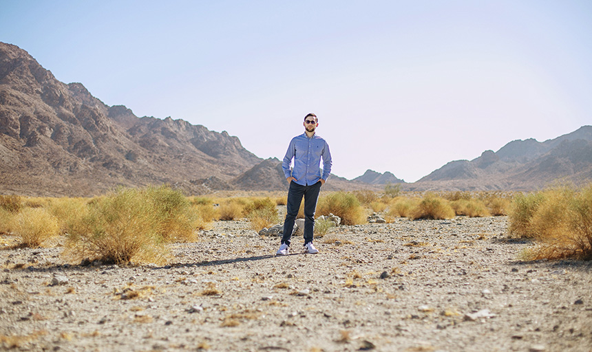 Young man standing in the center of a desert landscape with scrubby bushes around him and a pale blue sky