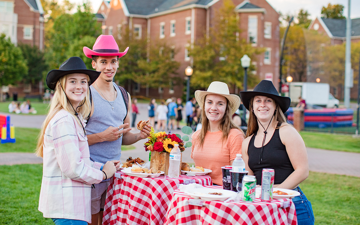 Students in cowboy hats stand around a table topped with a red-and-white checkered cloth, and they have plates of food in front of them.