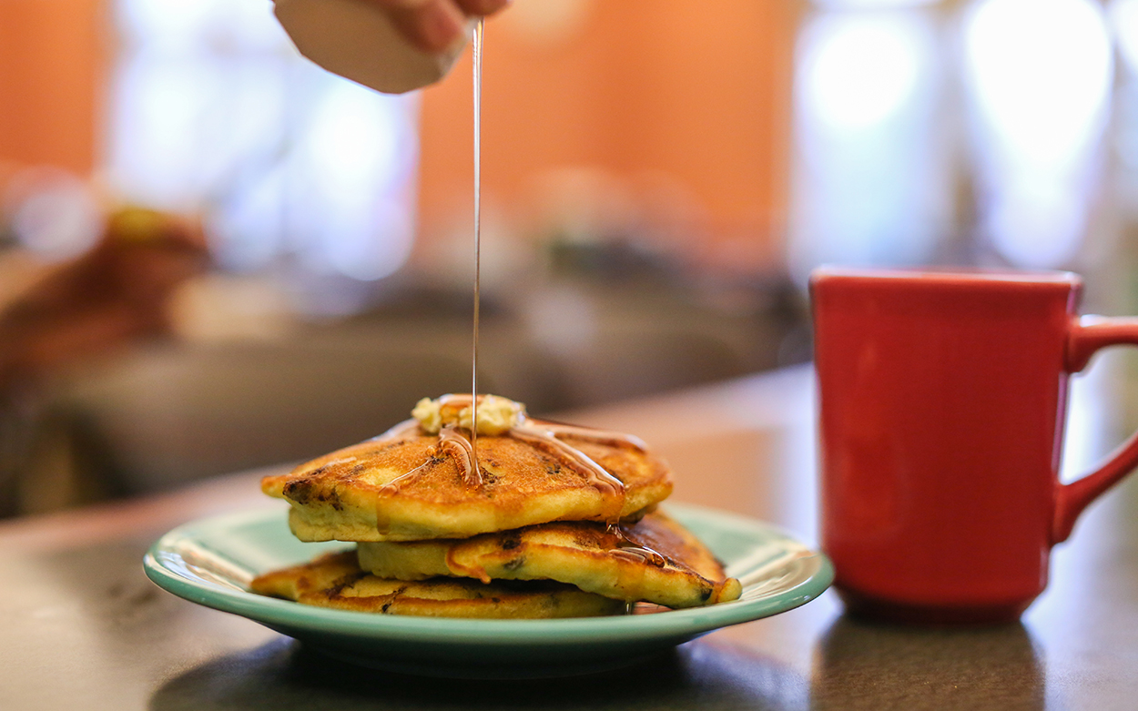 Syrup is drizzled on a plate of pancakes next to a red coffee mug on a table