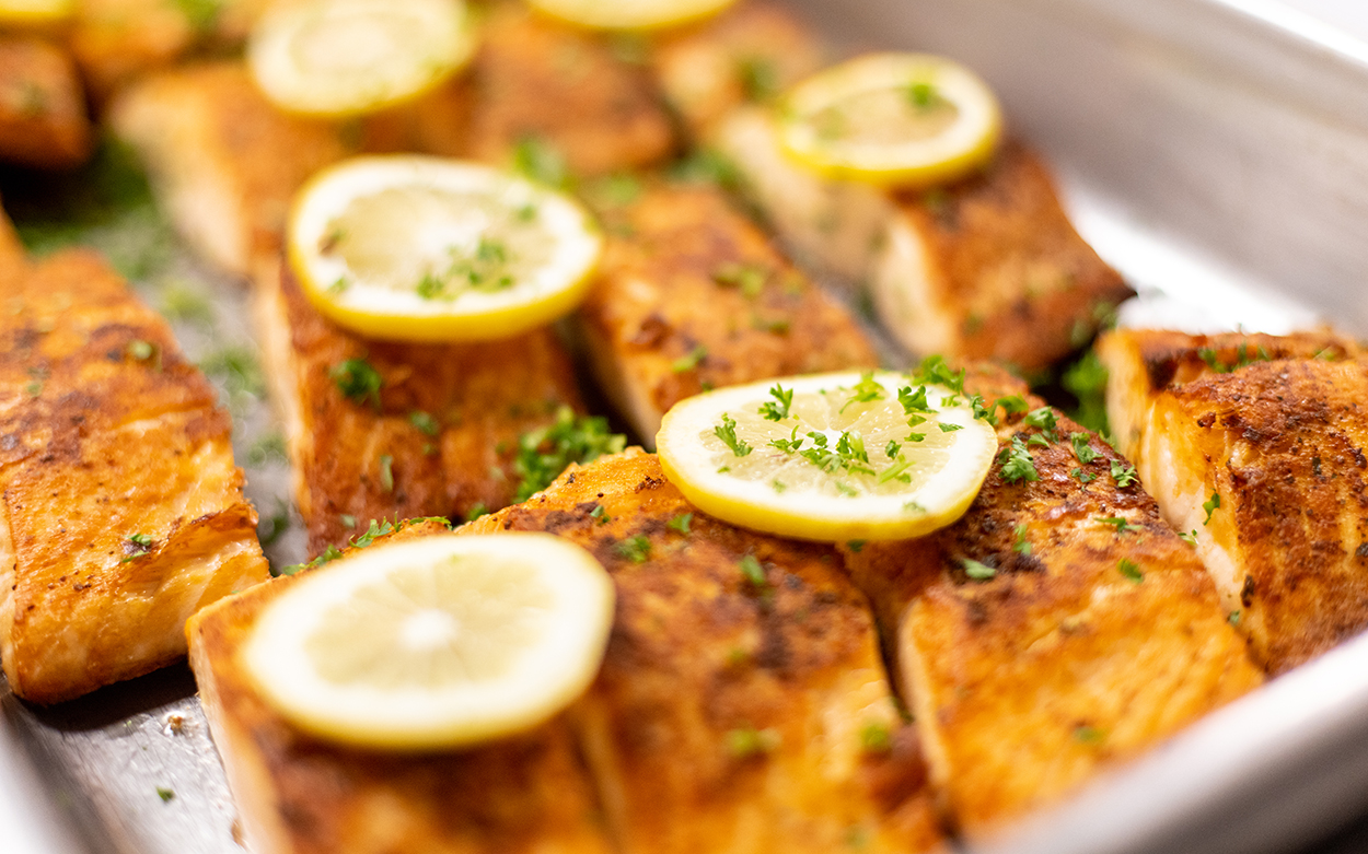 A tray of golden brown salmon filets, each with a slice of lemon and some chopped parsley on top.
