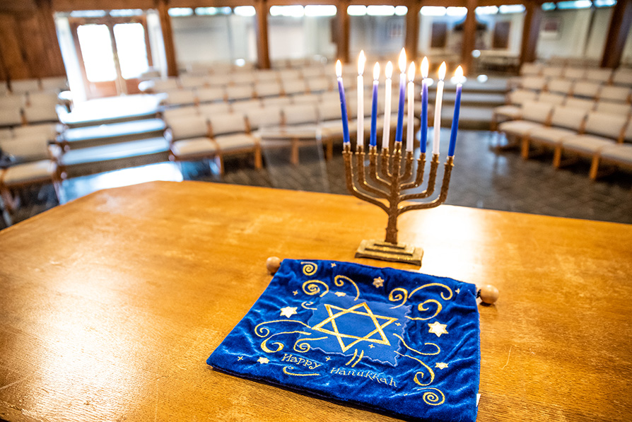 A menorah with all candles lit in the foreground sitting on a table with a blue cloth that has the Star of David embroidered in gold on it. Chapel pews in the background.