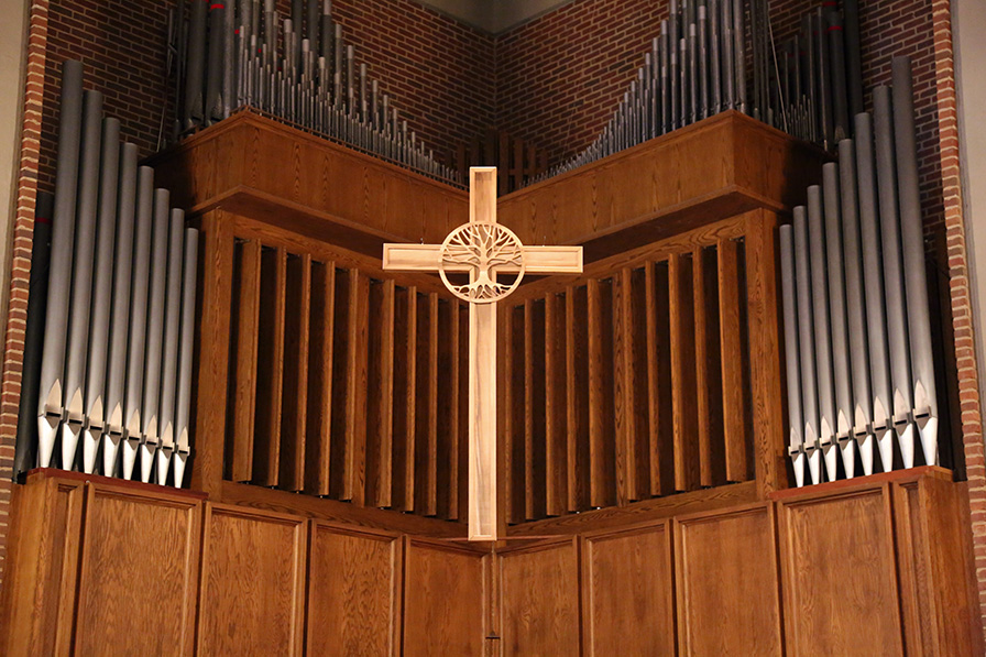 A large wooden cross in the center with the tree of life symbol carved into it and pipe organ in the background.