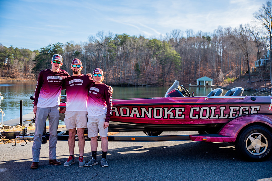 Three members of the bass fishing team, all male students in maroon jerseys, stand next to the Roanoke College bass fishing boat, which is painted maroon with a snazzy logo on the side.