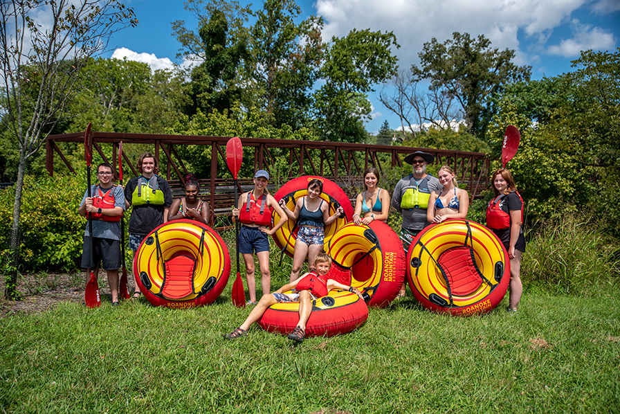A group of students stands next to the river, posing with each one holding a bright red and yellow inner tube for floating down the river.