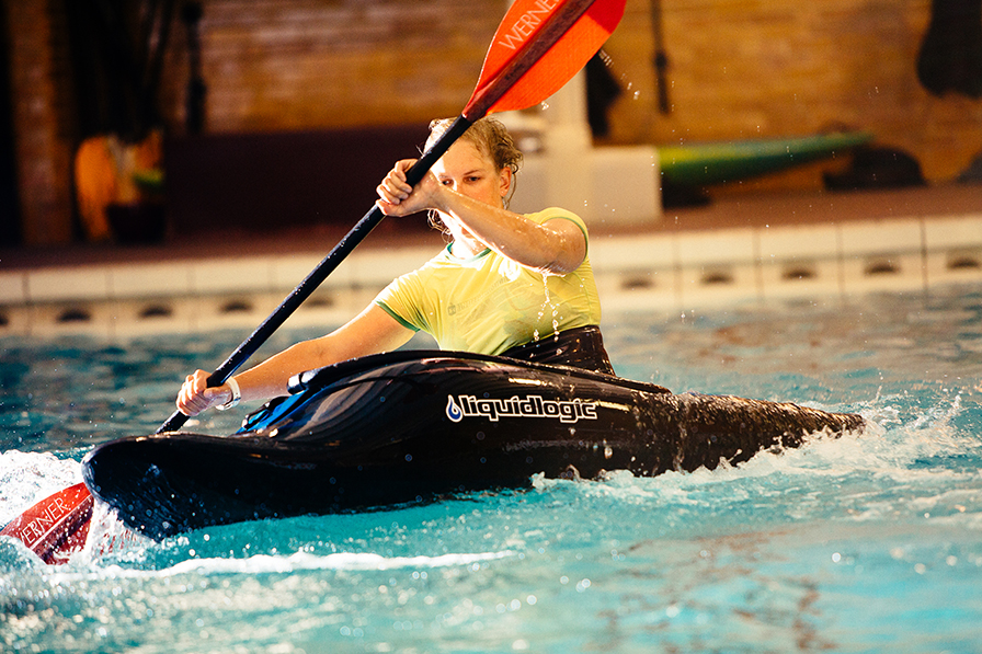 A young lady in a yellow shirt sits in a kayak i the swimming pool, where she is learning how to roll the boat.